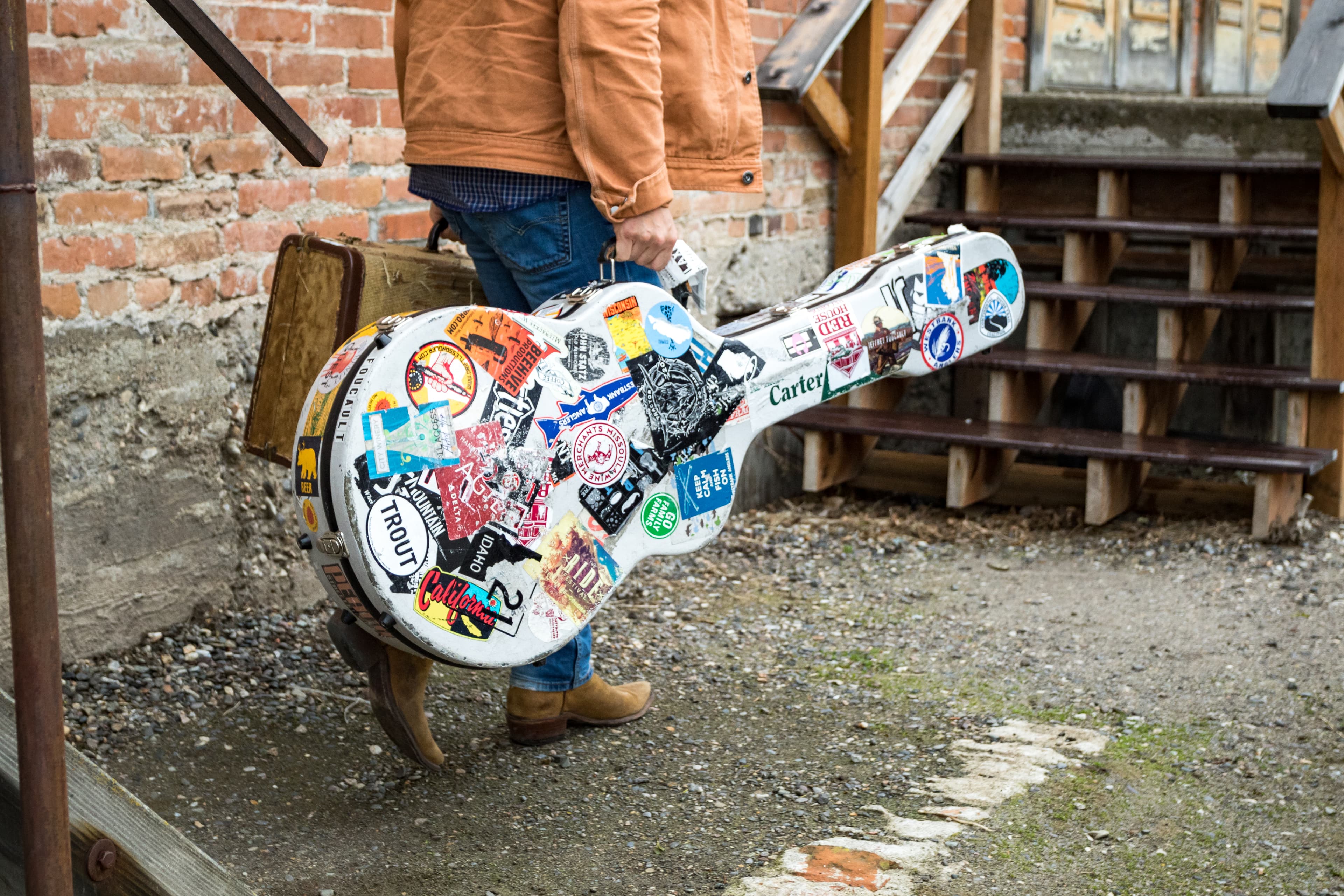 Child holding colorful sticker-covered guitar against brick wall.