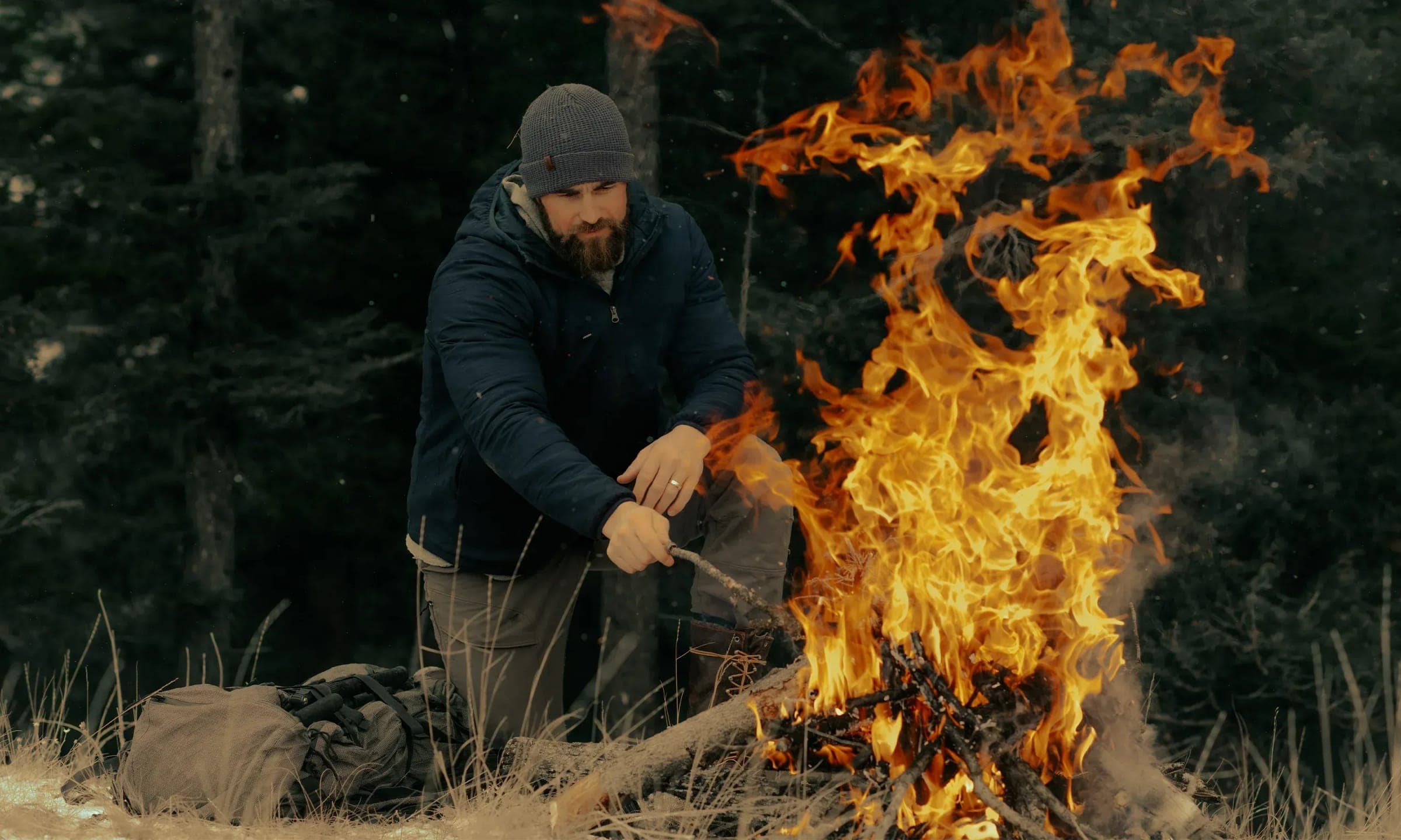 Man tending campfire in winter clothing and beanie outdoors.