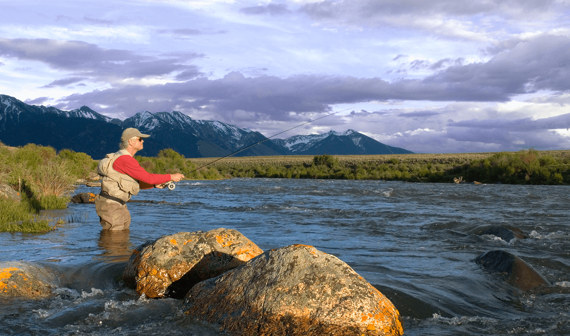 Fly fisherman casting in Madison River with Montana mountains in background.