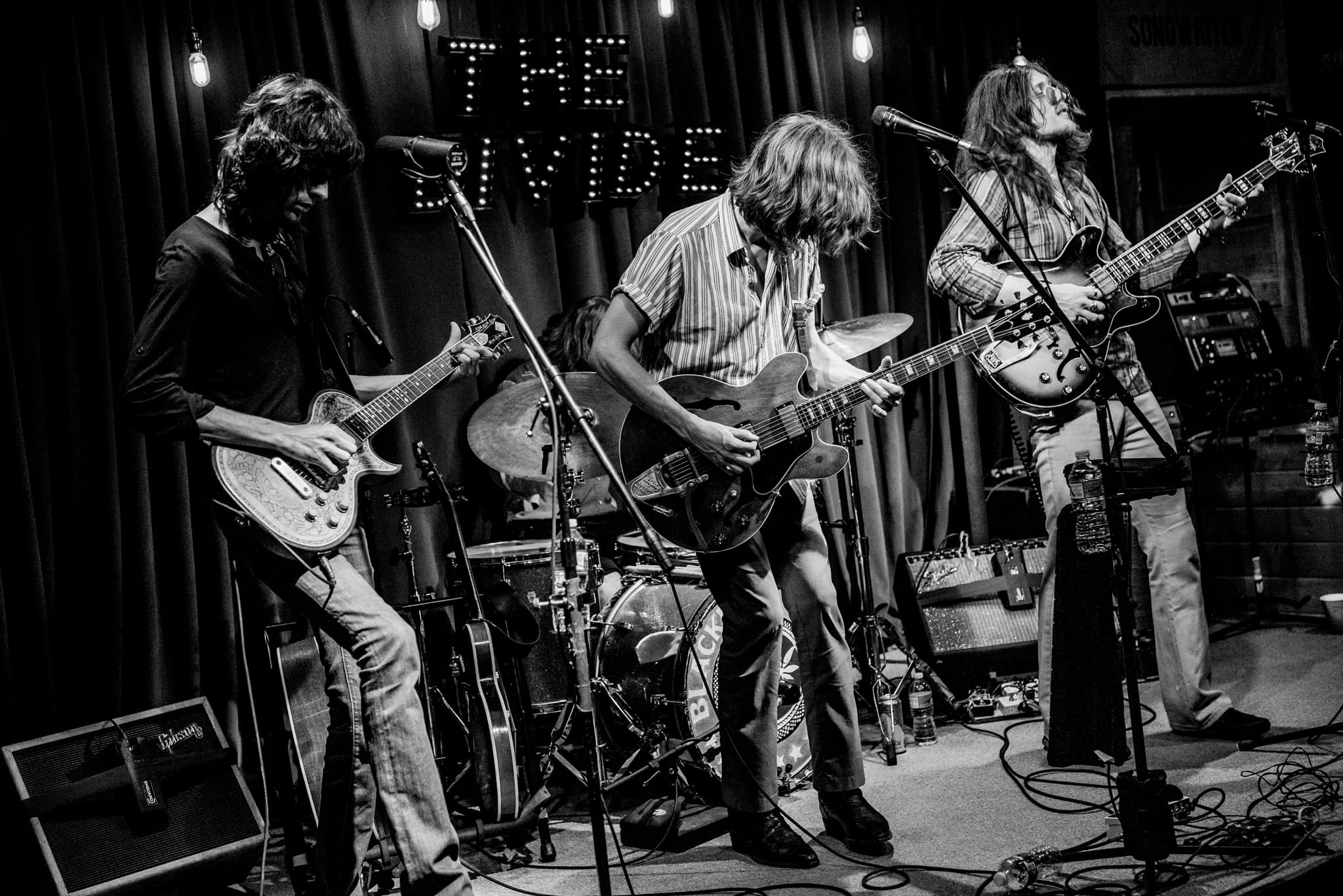 Three musicians performing on stage with guitars and drums in black and white.