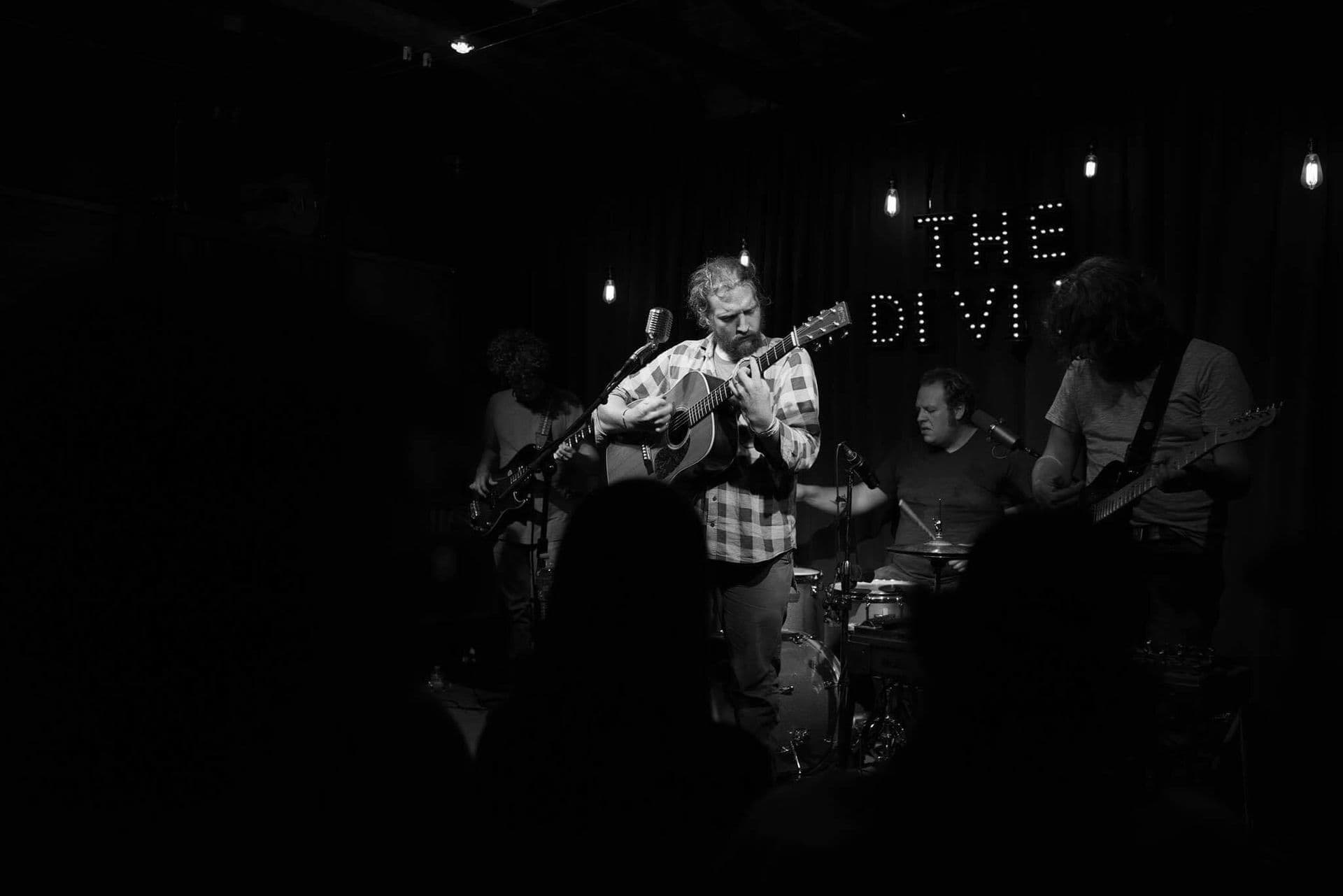 Black and white photo of musician performing with acoustic guitar on stage.