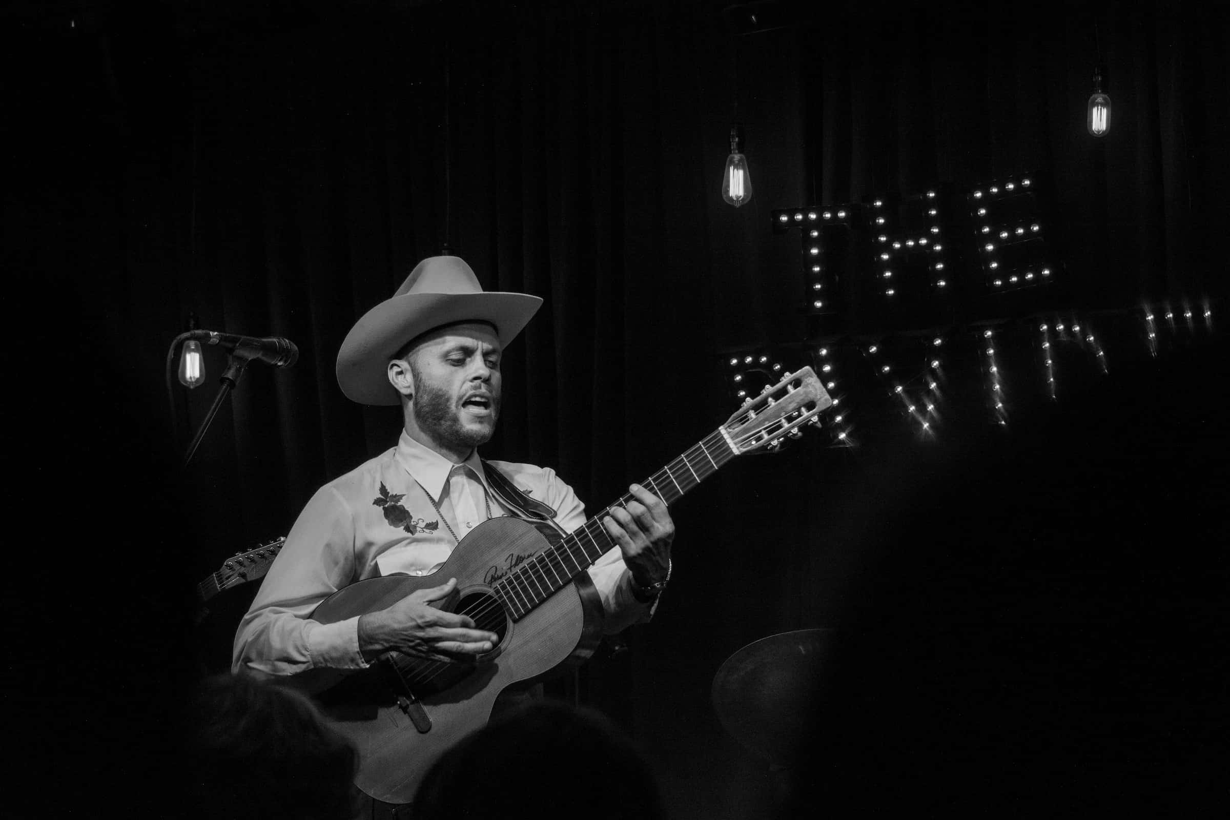 Charley Crockett performing with acoustic guitar on stage in black and white.